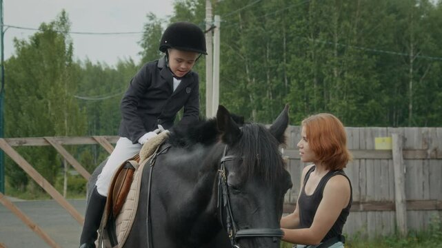 Young female horse riding coach backing up jockey boy with Down syndrome wearing helmet, fitted riding jacket, breeches, and tall boots mounting a black mare getting ready for ride