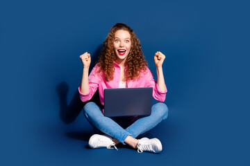 Full size photo of nice young woman sit floor raise fists laptop wear shirt isolated on dark blue color background