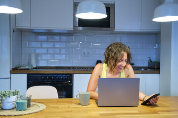 45 year old woman sitting at her kitchen table smiling using her laptop and smart phone