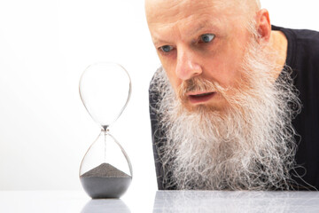 Elderly man with a thick beard gazes in awe at an hourglass, symbolizing the fleeting nature of time. His intense stare and the serene backdrop create a thought image on the inevitability of time