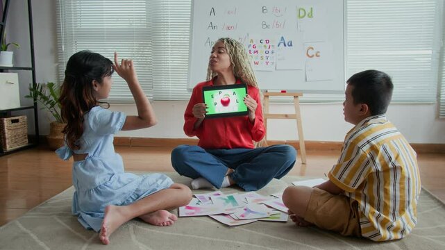 Long shot of Hispanic female tutor showing educational materials on digital tablet to two diverse schoolkids during speech therapy session