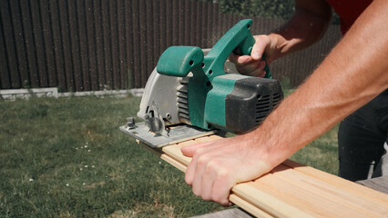 Carpenter sawing wood with handsaw. Male carpenter using electric circular saw. Wood chips flying. Unrecognizable worker cutting plank with circular saw at construction site.