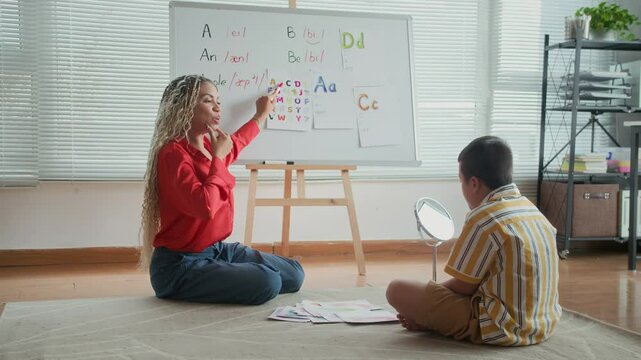 Long shot of teenage schoolboy sitting on floor in front of mirror and looking at female speech therapist teaching him to pronounce letters correctly at classroom