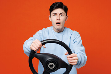 Young shocked scared mad sad man he wears blue sweatshirt casual clothes hold steering wheel driving car look camera isolated on plain red orange color background studio portrait. Lifestyle concept.