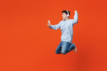 Full body side view young man in blue sweatshirt casual clothes jump high hold in hand use mobile cell phone listen music in headphones do winner gesture isolated on plain red orange color background.