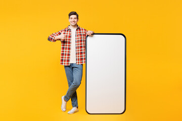 Full body young happy man he wears red checkered shirt casual clothes big huge blank screen mobile cell phone smartphone with area show thumb up isolated on plain yellow background. Lifestyle concept.