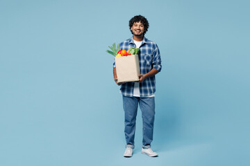 Full body young smiling happy Indian man wear shirt hold brown craft bag for takeaway mock up with food products look camera isolated on plain blue background Delivery service from shop or restaurant