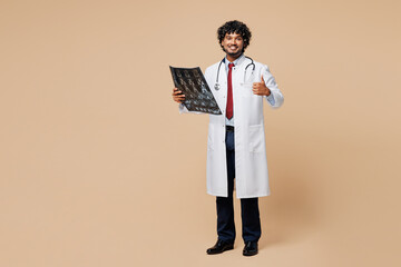 Young happy Indian doctor man wear white medical gown suit work in hospital clinic hold x-ray radiographic image ct scan head brain mri isolated on plain beige background. Healthcare medicine concept.