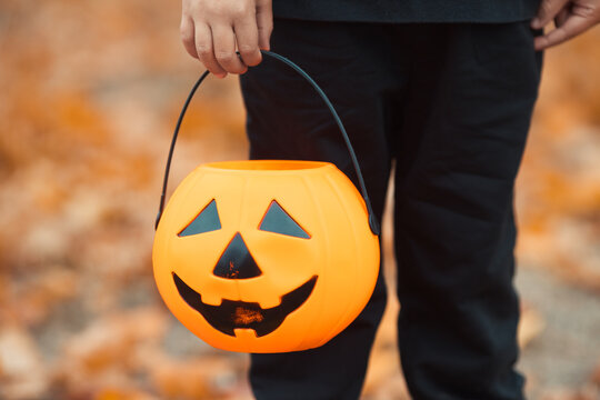 Close-up kid's hand hold a halloween candy bucket in pumpkin shape for trick or treat game. Celebrate all saints day with jack lantern.