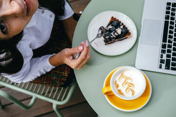 Top view of a young woman in a cafe eating cake, drinking coffee and working on a laptop.