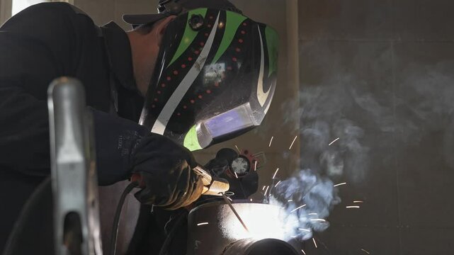 A welder, donning a protective helmet, engages in welding a metal pipe in a smoke-filled workshop, illustrating a scene of intense manual labor and craftsmanship.