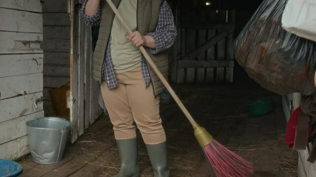 Tilt down of young female farm worker with Down syndrome wearing comfortable warm clothes and rubber boots brooming the straw-covered floor of equine stable on farm