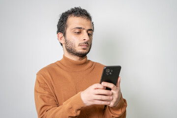 Young man in a brown sweater thoughtfully looking at his smartphone, isolated on a light background.