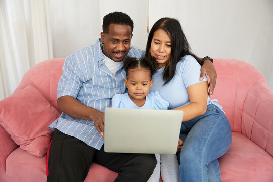 African American father and asian mother with daughter using laptop computer for watching movie or browsing internet on sofa