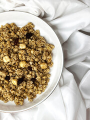 Granola with nuts and raisins in a white plate close-up on a white textile background. Top view. Healthy proper breakfast.