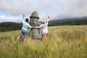 Jeju-si, Jeju-do, South Korea - May 28, 2018: Summer view of a couple holding and raising hands with Dolhareubang Stone at barley field against Halla Mountain in the background