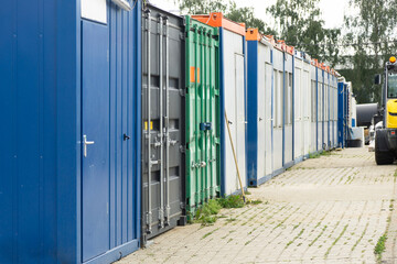 Row of industrial storage containers and portable site offices aligned along a paved walkway at a construction or logistics area, showing modular buildings and container units used for on-site work 