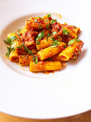 A close-up of Rigatoni pasta served with rich tomato sauce, garnished with fresh parsley. The dish is presented on a white plate, highlighting the vibrant colors and appetizing texture of the pasta.