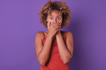 A young woman with curly hair stands against a vibrant purple backdrop, covering her mouth in...