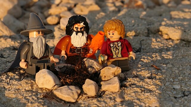 LEGO Hobit figures of Bilbo with The Ring, dwarf Bifur with LEGO fish and wizard Gandalf talking around non lit fireplace on shoreline rocks, summer evening sunshine. 