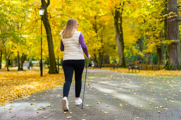 Obraz premium Blonde woman dressed sporty exercising nordic walking on alley full of yellow fallen leaves in park on autumn day. Back view. Autumnal training of Nordic walking.