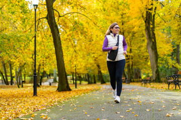 Pretty middle-aged blonde woman dressed sporty running on alley full of beautiful autumnal trees in city park on autumn day. Front view. Autumnal training. Healthy running.