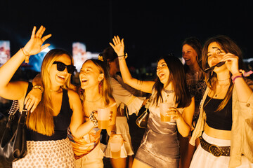 Group of young women dancing and enjoying their drinks at an outdoor music festival at night.