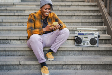 Trendy curvy african woman sitting on city stairs while using mobile phone and listening music with vintage boombox stereo - Urban lifestyle concept