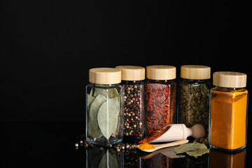 Different spices in glass jars on black mirror surface, space for text