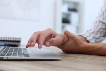 Man suffering from pain in wrist while working on laptop at table indoors, closeup. Carpal tunnel...