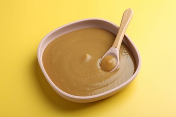 Delicious baby food with spoon in bowl on yellow table