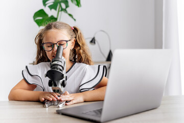 A young schoolgirl uses a microscope and a laptop for her studies. Perfect for content on education, science, and technology, highlighting children's curiosity and learning in the digital age.