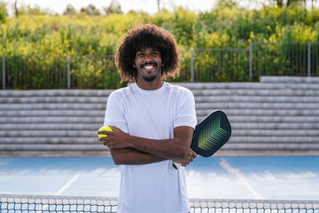 Smiling African American pickleball player holding paddle and ball, outdoor court © Koldo_Studio