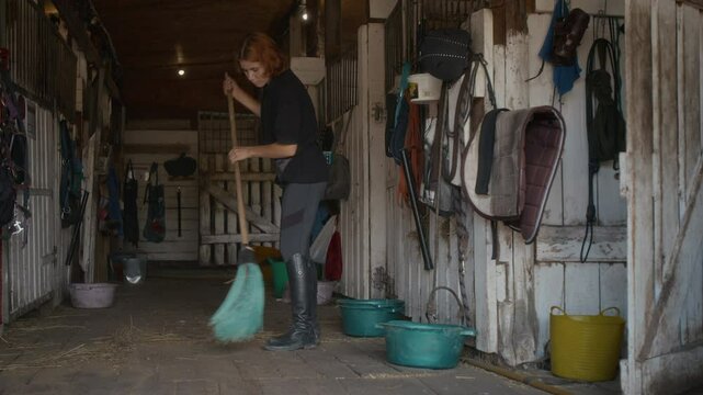 Wide shot of young ginger-haired Caucasian horse rider wearing casual comfortable clothes and tall black boots holding broom and cleaning the straw-covered floor in stable on farm