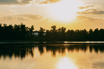 Fototapeta premium A bright light orange sunset on a calm river with a bank lined with tall trees. The rays of the luminary glare and reflect from the water surface. Sunny evening in nature.