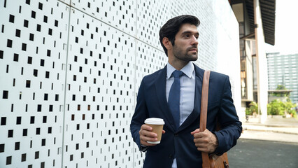 Fototapeta premium Skilled business man walking in suit outfit while holding coffee cup. Professional manager standing near architectural building. Going to working in morning, seeking for successful job. Exultant.