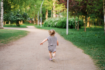 Cute Caucasian toddler child 2-years-old girl walks in a green park at sunset in summer. The concept of a joyful childhood and active pastime.