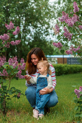 A charming child girl toddler and a young mother against a background of lilacs in a green summer park. Active childhood.