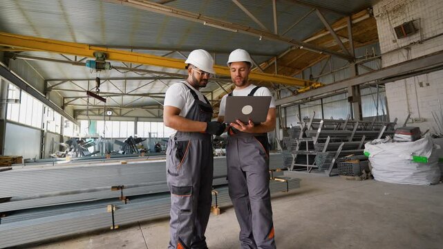 Caucasian foreman and Indian worker with laptop in warehouse metal profile