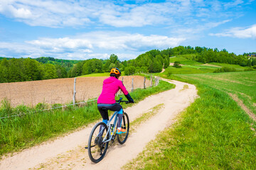 Naklejka premium Woman cyclist riding bicycle on gravel countryside road along green meadows and hills, Suwalski Landscape Park, Podlasie, Poland