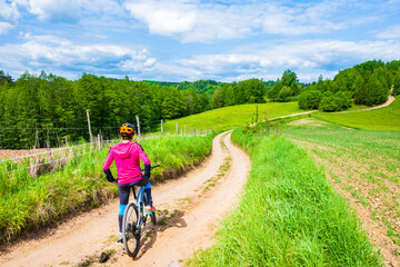 Obraz premium Woman cyclist riding bicycle on gravel countryside road along green meadows and hills, Suwalski Landscape Park, Podlasie, Poland