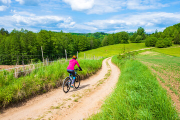 Woman cyclist riding bicycle on gravel countryside road along green meadows and hills, Suwalski...