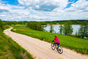 Woman cyclist riding bicycle on gravel countryside road along lake and green meadows, Suwalski Landscape Park, Podlasie, Poland