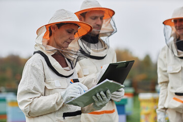 Side view of serious middle aged female apiarist in protective bee suit writing in notepad while keeping beehives with colleagues at apiary farm, copy space