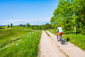 Naklejka premium Woman cyclist riding bicycle on gravel rural road among green meadows and fields, Suwalski Landscape Park, Podlasie, Poland