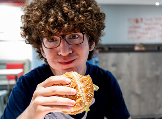 Nice portrait of a young caucasian man with curly hair and glasses smiling looking into the camera, he is eating a sandwich in a fast food restaurant. Young lifestyle and fast food consumption.