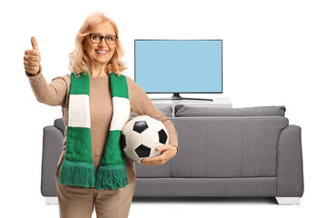 Middle aged female football fun holding a ball and gesturing thumbs up in front of tv