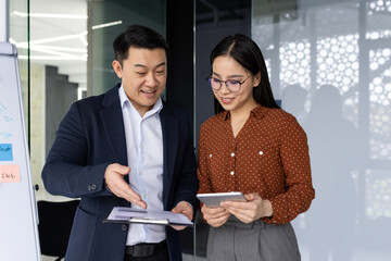 Asian business professionals engaged in collaborative discussion in modern office environment. Man and woman review documents and tablet, demonstrating teamwork and technology use.