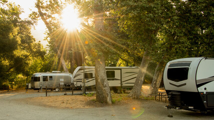 Rv camping trailers parked at campsite under trees with sunburst coming through 