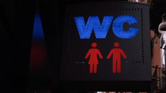 A WC sign hanging on the street, indicating a public restroom, with flashing blue letters and red icons, framed by red string lights and alternating LEDs. Dark setting. Handheld closeup shot.
